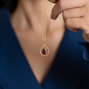 May include: A gold-colored necklace with a teardrop pendant. The pendant features a deep red gemstone surrounded by a gold-colored, twisted rope-like frame. The necklace is held up against a dark blue background.