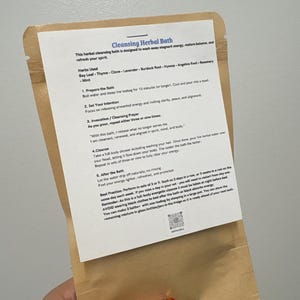 May include: A brown paper packet with a white label detailing a cleansing herbal bath. The label lists herbs and instructions, including preparing the bath, setting intentions, and cleansing. The packet is held by a hand with red nail varnish.