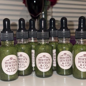 Puede incluir: Una colección de frascos cuentagotas de vidrio llenos de un líquido verde brillante. Cada frasco tiene una tapa cuentagotas negra y una etiqueta blanca con el texto "Mirth's Jewels & Co.". Los frascos están dispuestos en fila.