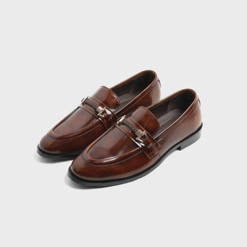 May include: A pair of brown leather loafers with a glossy finish. Each shoe features a decorative strap with a metal buckle. The shoes have a classic, slip-on design, suitable for formal or semi-formal occasions.