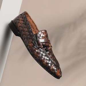 May include: A single brown leather loafer with a woven design. The shoe features a rounded toe, a small tassel detail, and a dark sole. The leather has a polished finish, and the shoe is set against a neutral background.