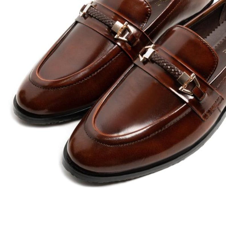 May include: A pair of brown leather loafers with a glossy finish. Each shoe features a decorative strap with a woven detail and a gold-colored buckle. The shoes have a classic design with a rounded toe and a black sole.