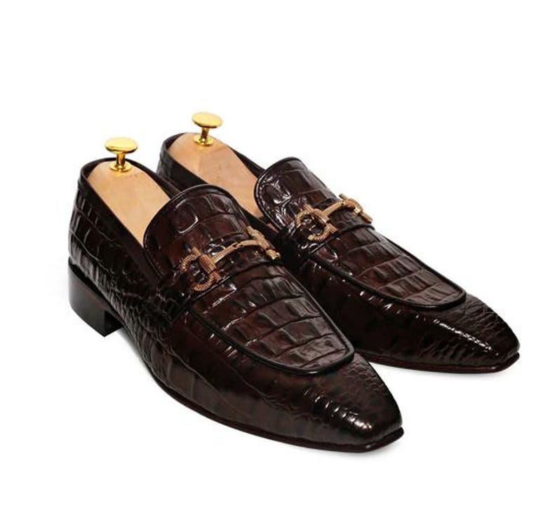 May include: A pair of brown leather loafers with a crocodile pattern. The loafers have gold buckles and a pointed toe.