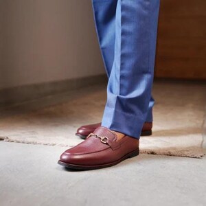May include: Burgundy leather loafers with a gold-colored metal buckle detail. The shoes are worn with blue trousers. The loafers have a classic, slip-on design and a polished finish.