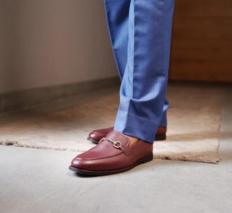 May include: Burgundy leather loafers with a gold-colored metal buckle detail. The shoes are worn with blue trousers. The loafers have a classic, slip-on design and a polished finish.