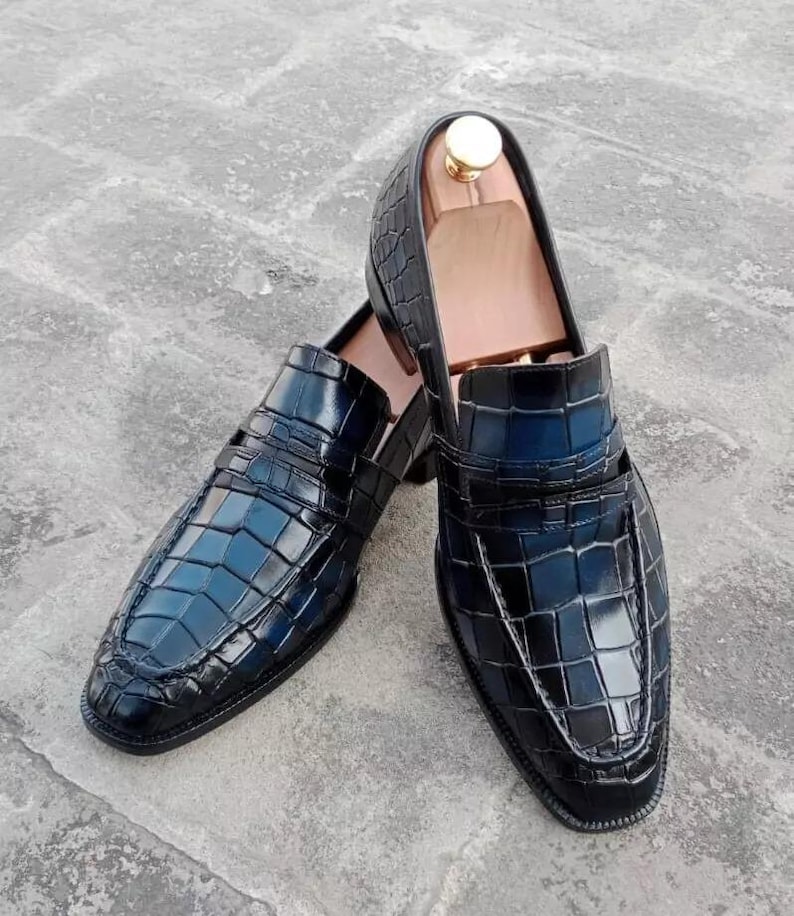 Puede incluir: Un par de mocasines de cuero negro con estampado de cocodrilo. Los mocasines tienen punta puntiaguda y un dise&ntilde;o de deslizamiento.