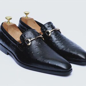 May include: A pair of black leather loafers with a textured surface and gold-colored metal accents. The shoes have a classic design with a slip-on style and are displayed with wooden shoe trees inside.