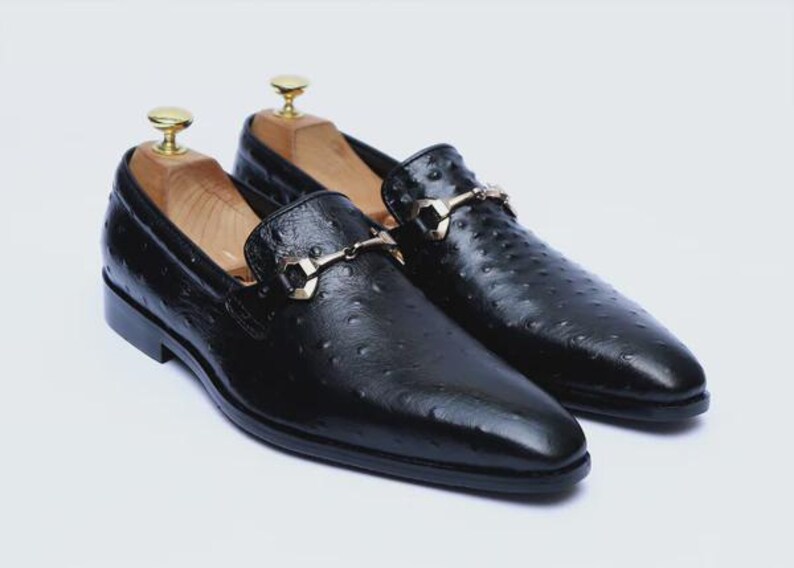 May include: A pair of black leather loafers with a textured surface and gold-colored metal accents. The shoes have a classic design with a slip-on style and are displayed with wooden shoe trees inside.