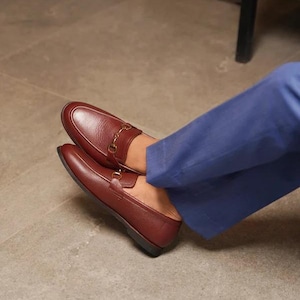 May include: A pair of burgundy leather loafers with gold-colored metal accents. The shoes are worn with blue pants. The loafers have a classic design, suitable for formal or casual wear.