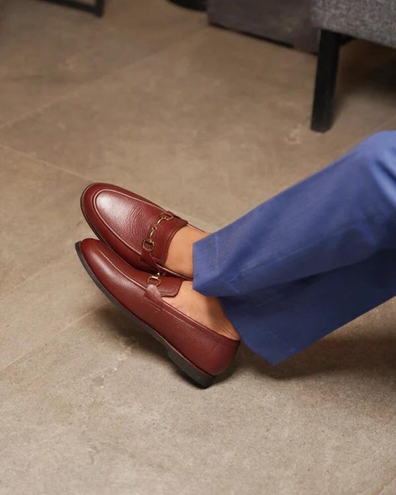May include: A pair of burgundy leather loafers with gold-colored metal accents. The shoes are worn with blue pants. The loafers have a classic design, suitable for formal or casual wear.
