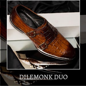 May include: Brown leather double monk strap shoe with a crocodile texture. The shoe features two gold-colored buckles and a sleek design. The text "DILEMONK DUO" is displayed at the bottom of the image.