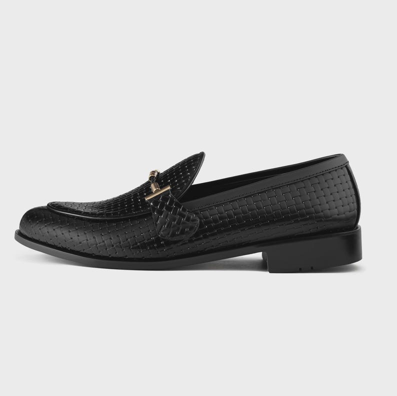 May include: Black leather loafer with a woven texture and a gold-colored buckle. The shoe has a low heel and a classic design, suitable for formal or semi-formal occasions.