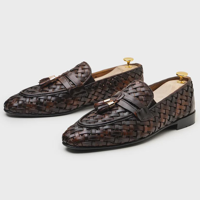 May include: A pair of brown woven leather loafers with tassel accents. The shoes have a classic design with a woven pattern and a leather strap across the top. The loafers are displayed on a light background.