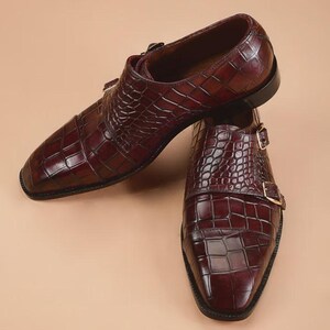 May include: A pair of burgundy leather dress shoes with a crocodile skin texture. The shoes feature a double monk strap design with gold-colored buckles and a square toe. The shoes are displayed on a neutral background.