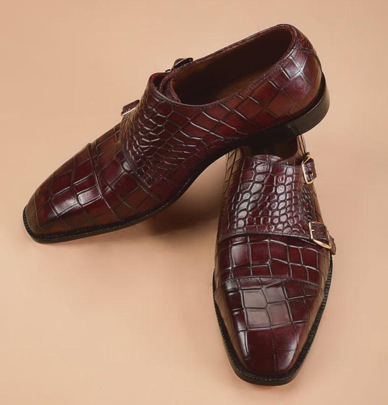 May include: A pair of burgundy leather dress shoes with a crocodile skin texture. The shoes feature a double monk strap design with gold-colored buckles and a square toe. The shoes are displayed on a neutral background.