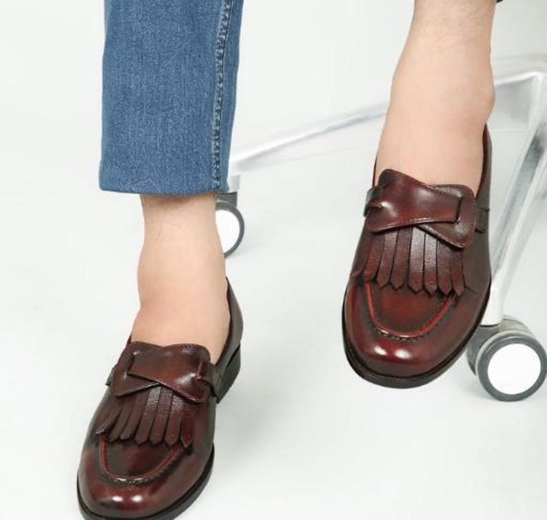 May include: A pair of brown leather loafers with fringe detailing. The loafers have a pointed toe and a low heel.
