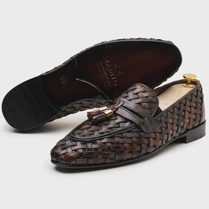 May include: Brown leather woven loafers with a tassel detail and a leather sole. The shoes have a classic design and are suitable for formal or casual wear. The sole of the shoe in the background has the text "MARITO MADE IN ITALY".