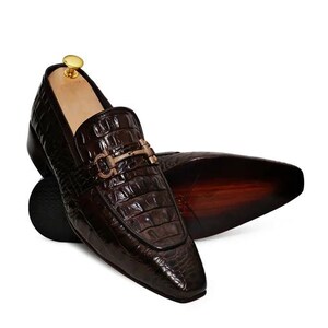 May include: Brown leather loafers with a gold buckle detail. The loafers have a crocodile-embossed pattern and a pointed toe.