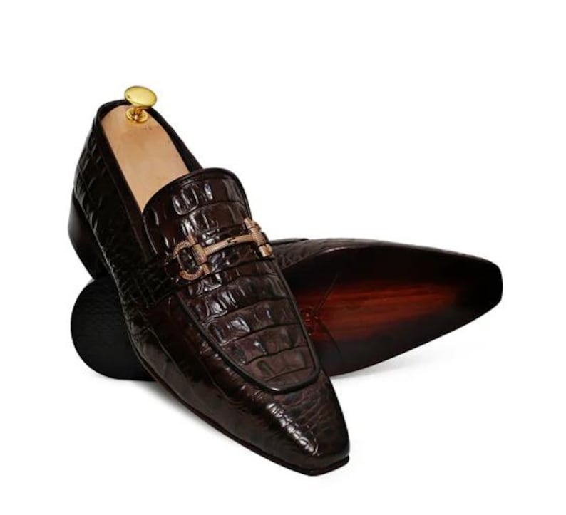 May include: Brown leather loafers with a gold buckle detail. The loafers have a crocodile-embossed pattern and a pointed toe.
