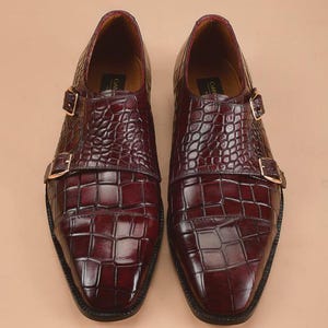 May include: A pair of burgundy leather double monk strap shoes. The shoes feature a textured, crocodile-like pattern and gold-colored buckles. The soles are black, and the shoes have a polished finish, suitable for formal occasions.