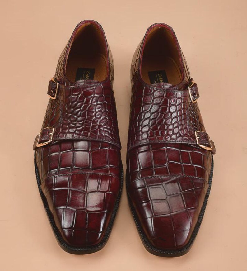 May include: A pair of burgundy leather double monk strap shoes. The shoes feature a textured, crocodile-like pattern and gold-colored buckles. The soles are black, and the shoes have a polished finish, suitable for formal occasions.