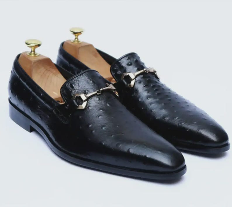 May include: A pair of black leather loafers with a textured finish and gold-colored metal accents. The shoes have a classic slip-on design and are displayed on wooden shoe trees.