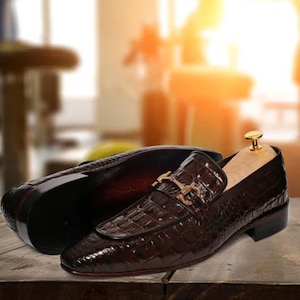May include: A pair of brown leather loafers with a crocodile pattern. The loafers have a gold buckle and a wooden shoe tree inside.