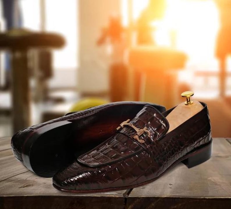 May include: A pair of brown leather loafers with a crocodile pattern. The loafers have a gold buckle and a wooden shoe tree inside.