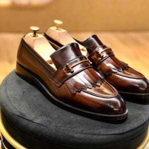 以下が含まれることがあります： 茶色の革製ローファーのペア。装飾的なフリンジと金色の金具が特徴です。靴は黒い丸い表面に展示されており、洗練された仕上げとクラシックなデザインを際立たせています。ローファーは洗練されたモダンなシルエットをしています。