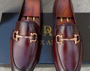 Könnte beinhalten: Ein Paar polierte, weinrote Leder-Loafer mit goldfarbenen Beschlägen. Die Schuhe haben ein klassisches Design mit runder Spitze und einem dekorativen Riemen. Holzschuhspanner sind eingesetzt. Auf einer schwarzen Box mit dem Text "DA X AI".