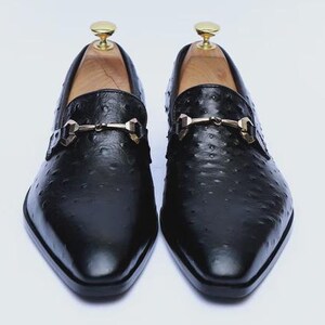May include: A pair of black leather loafers with a textured finish. Each shoe features a gold-colored metal buckle and a wooden shoe tree. The shoes have a pointed toe and are displayed on a white background.