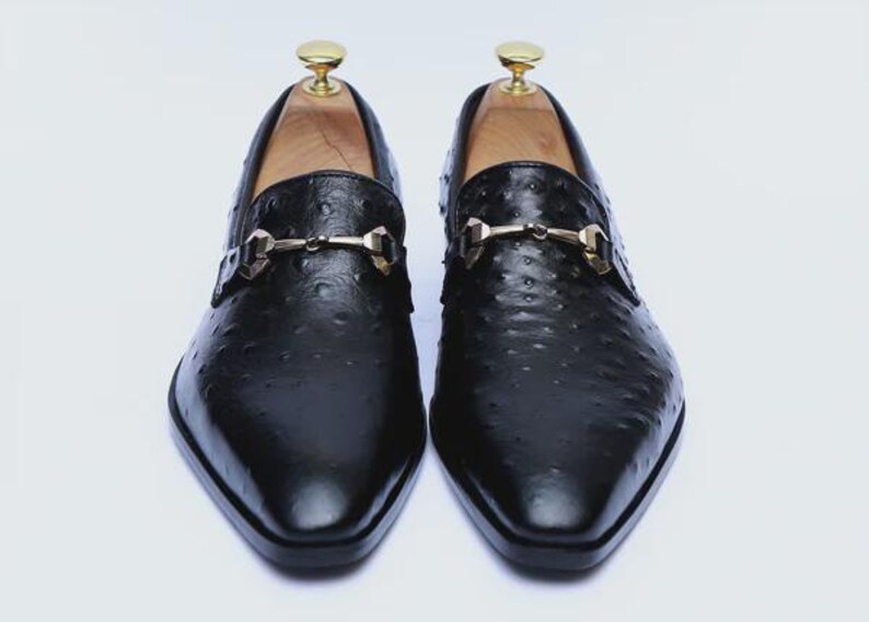 May include: A pair of black leather loafers with a textured finish. Each shoe features a gold-colored metal buckle and a wooden shoe tree. The shoes have a pointed toe and are displayed on a white background.