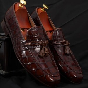 May include: A pair of brown leather loafers with a crocodile skin pattern. The shoes have a tassel detail and are displayed with wooden shoe trees inside. The loafers are a dark brown color and have a glossy finish.
