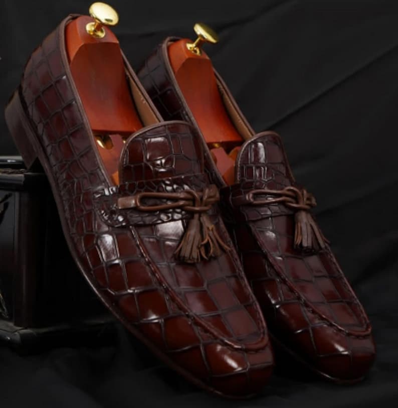 May include: A pair of brown leather loafers with a crocodile skin pattern. The shoes have a tassel detail and are displayed with wooden shoe trees inside. The loafers are a dark brown color and have a glossy finish.