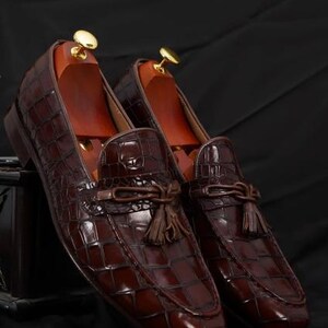 May include: A pair of brown leather loafers with a crocodile skin texture. The shoes have a tassel detail and are displayed with wooden shoe trees. The loafers are a rich, dark brown color.
