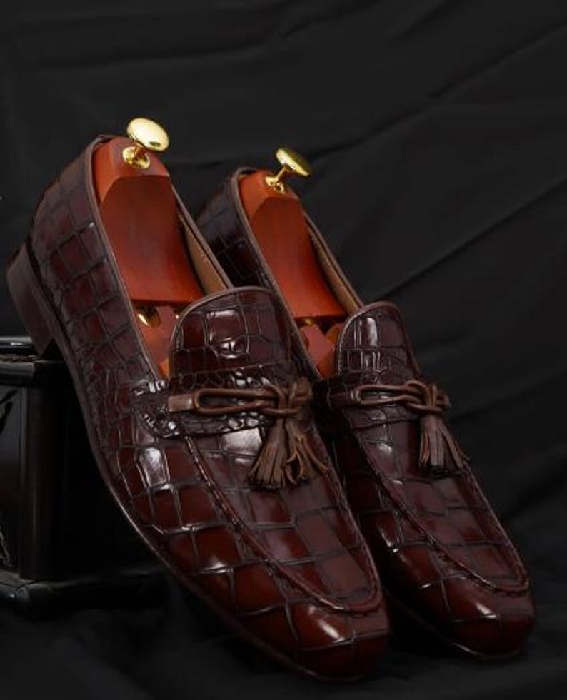 May include: A pair of brown leather loafers with a crocodile skin texture. The shoes have a tassel detail and are displayed with wooden shoe trees. The loafers are a rich, dark brown color.
