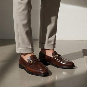 May include: A pair of brown leather loafers with gold-colored hardware. The shoes have a classic design with a polished finish. The image shows the shoes from a low angle, highlighting their shape and details. The loafers are worn with light-colored pants.