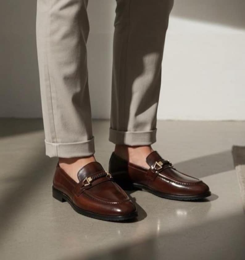 May include: A pair of brown leather loafers with gold-colored hardware. The shoes have a classic design with a polished finish. The image shows the shoes from a low angle, highlighting their shape and details. The loafers are worn with light-colored pants.