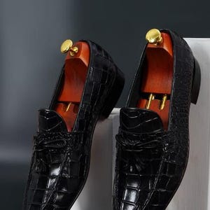 May include: A pair of black leather loafers with a crocodile skin texture. The shoes have a tassel detail on the vamp and are displayed with wooden shoe trees. The loafers are on a white surface against a dark background.