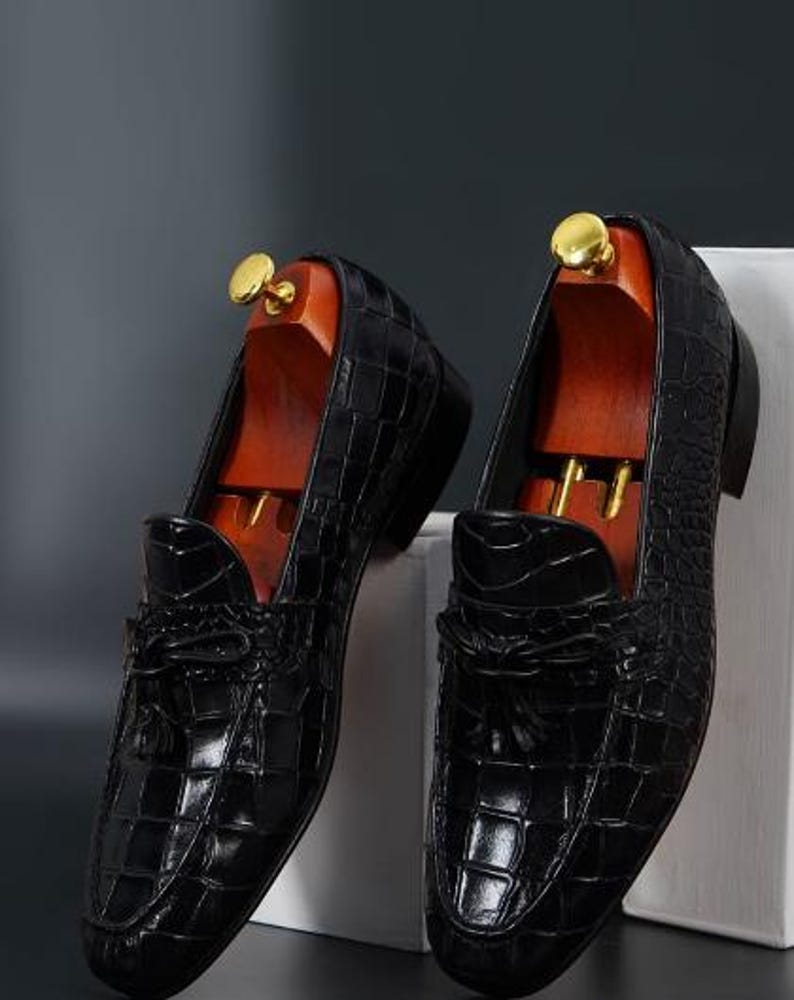 May include: A pair of black leather loafers with a crocodile skin texture. The shoes have a tassel detail on the vamp and are displayed with wooden shoe trees. The loafers are on a white surface against a dark background.