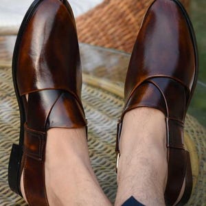 May include: Dark brown leather sandals with a polished finish. The sandals feature a closed-toe design, a strap across the top, and a buckle fastening. The shoes are a deep brown colour.
