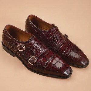 May include: A pair of burgundy leather double monk strap shoes. The shoes feature a textured, crocodile-like pattern and gold-colored buckles. The shoes have a square toe design and a polished finish.