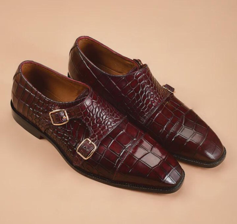 May include: A pair of burgundy leather double monk strap shoes. The shoes feature a textured, crocodile-like pattern and gold-colored buckles. The shoes have a square toe design and a polished finish.