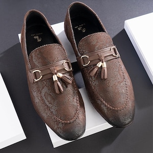 May include: A pair of brown leather loafers with a textured snakeskin pattern. The shoes feature a decorative strap with gold-colored hardware and small tassels. The loafers are displayed on a white surface against a dark background.