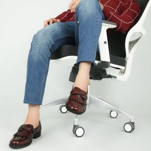 May include: A person sitting in a white office chair with a black seat. They are wearing blue jeans and brown leather loafers with fringe detailing.