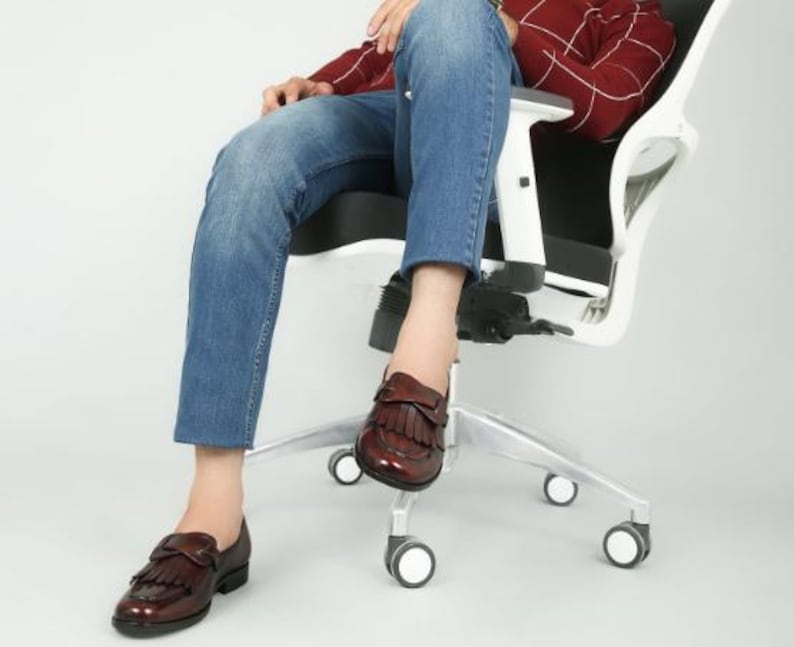 May include: A person sitting in a white office chair with a black seat. They are wearing blue jeans and brown leather loafers with fringe detailing.