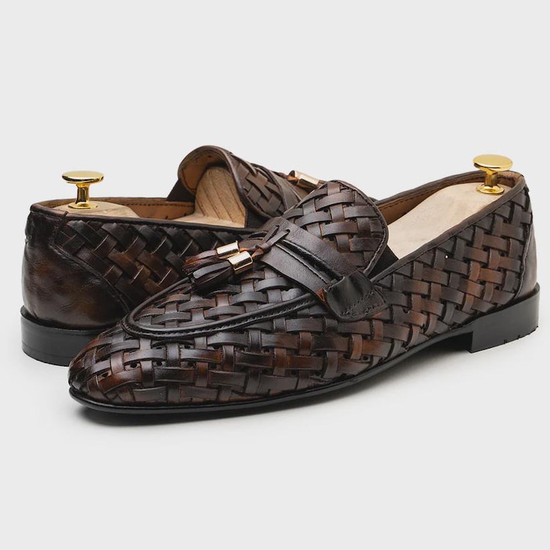 May include: A pair of brown leather loafers with a woven design. The shoes feature a tassel detail and a gold-colored accent on the top. The shoes are displayed on a light background.