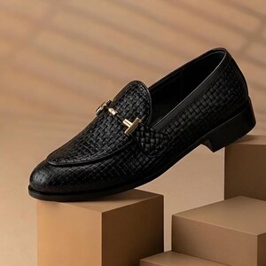 May include: A black leather loafer with a woven texture and a gold-colored buckle. The shoe is displayed on a series of brown blocks, showcasing its design and craftsmanship. The classic style is suitable for various occasions.