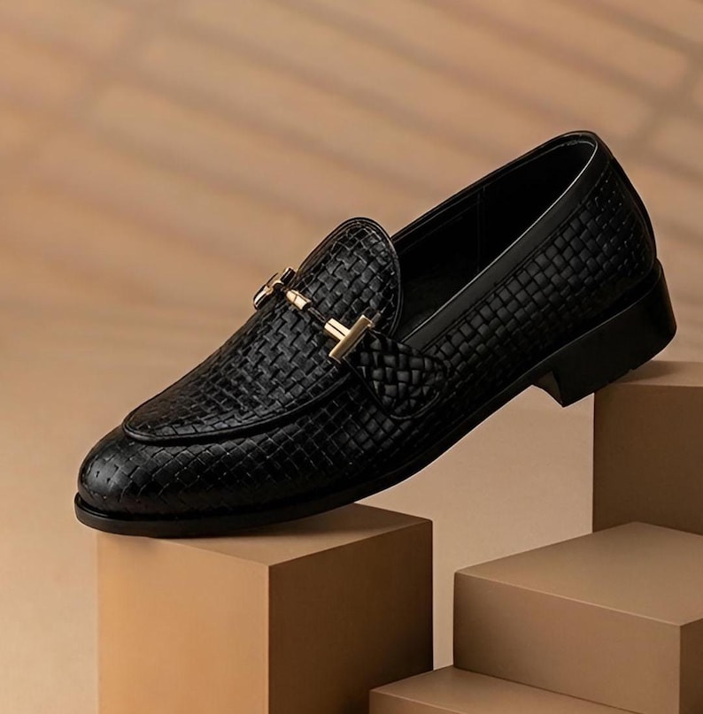 May include: A black leather loafer with a woven texture and a gold-colored buckle. The shoe is displayed on a series of brown blocks, showcasing its design and craftsmanship. The classic style is suitable for various occasions.