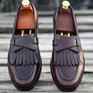 May include: A pair of burgundy leather tassel loafers with a fringe detail. The loafers are on a wooden surface.
